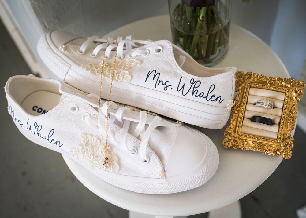 White Converse sneakers personalized with Mrs. Whalen and lace flower details, displayed on a table beside a gold framed wedding ring box.