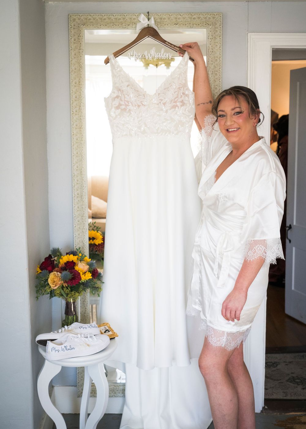 Smiling bride in a white satin robe holds up her lace wedding gown on a hanger beside sunflower bouquet and bridal garters.