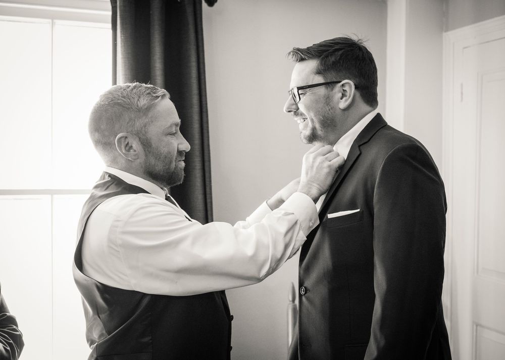 Black and white photo of a groomsman in a vest adjusting the tie of the groom in a dark suit before the wedding.