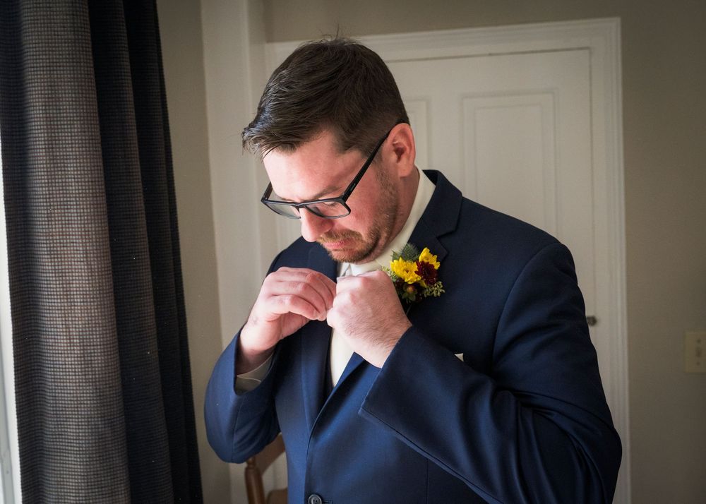 Groom in glasses and a navy suit adjusts his yellow flower boutonniere while getting ready for his wedding day.