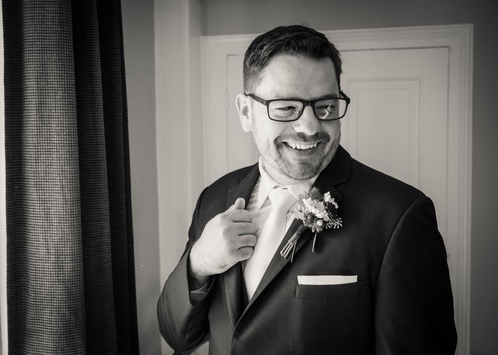 Black and white portrait of a smiling groom in glasses and dark suit adjusting his jacket, wearing a floral boutonniere.