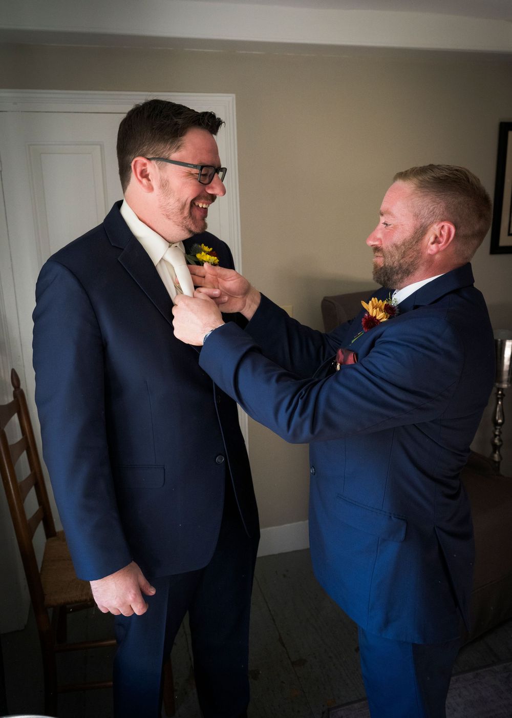 A smiling groom in a navy suit and glasses has a boutonniere pinned to his lapel by a groomsman in a blue suit.