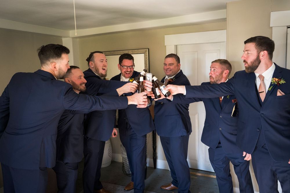 A group of groomsmen in navy suits gather in a close circle, laughing and toasting with drinks before the wedding.
