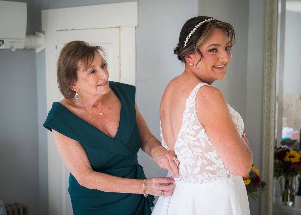 A woman in a green dress helps a smiling bride in a lace gown with a hair accessory fasten her wedding dress.