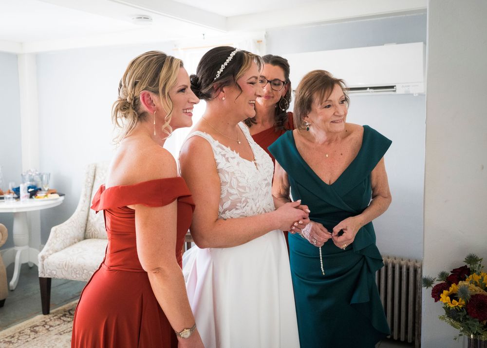 Bride in a white lace gown gets ready for her wedding with bridesmaids in rust and emerald dresses and her mother nearby.