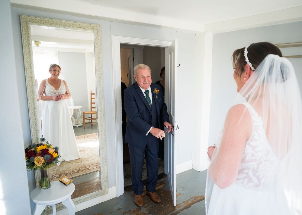 A father in a navy suit opens a door and sees his daughter in her wedding gown and veil for an emotional first look.