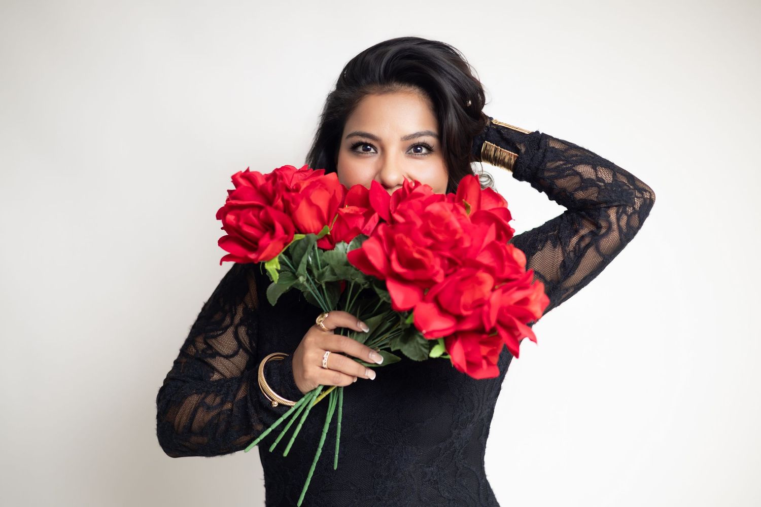 Woman in black lace dress holding bouquet of red roses covering her face, with dark hair against white background.