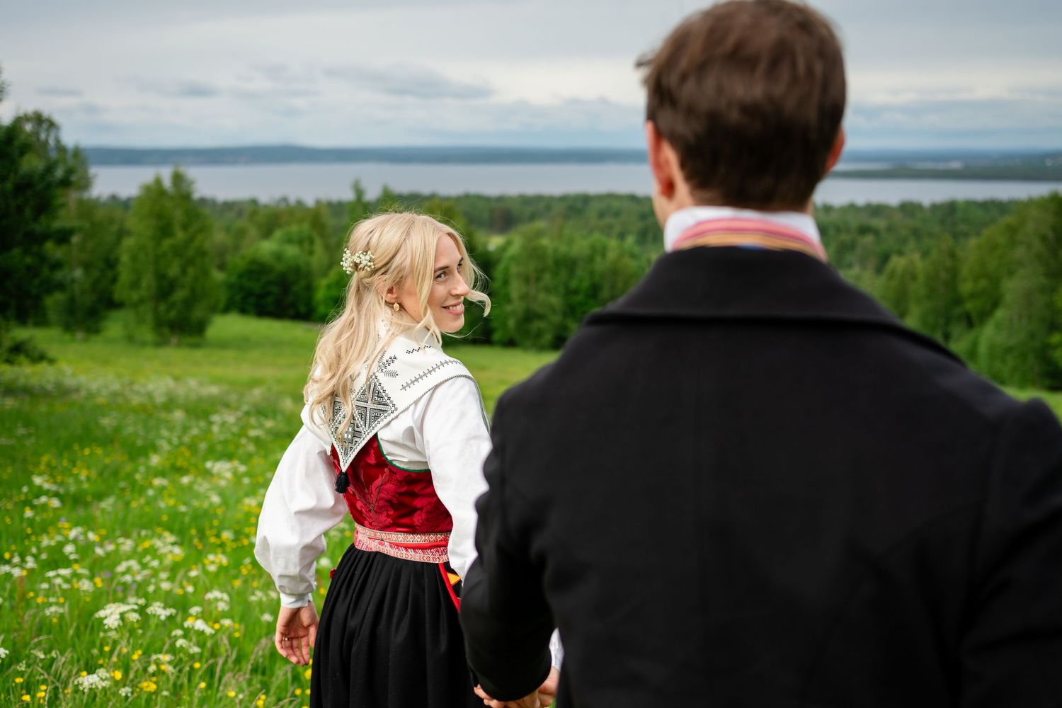 Couple in traditional Norwegian bunad attire holding hands in a green meadow with scenic fjord views.
