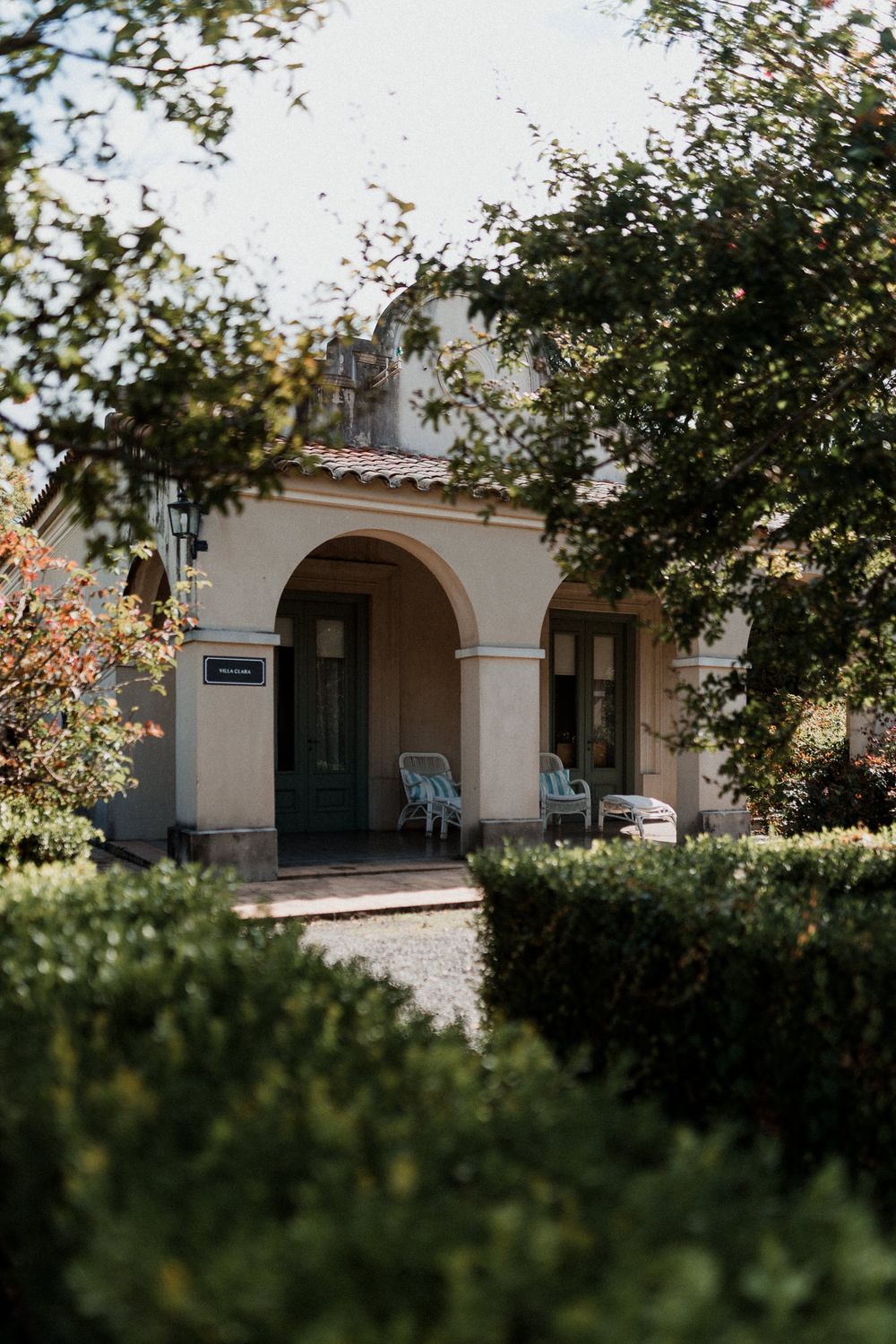 Charming Mediterranean-style home with arched porch, blue chairs, and lush garden surroundings on a sunny day.