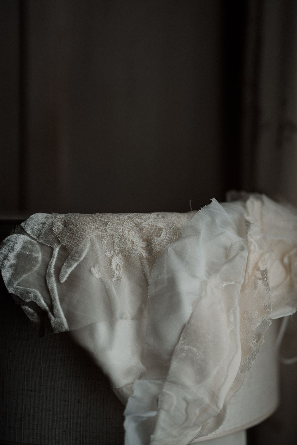 Delicate white lace and chiffon wedding dress draped over a chair in soft moody lighting.