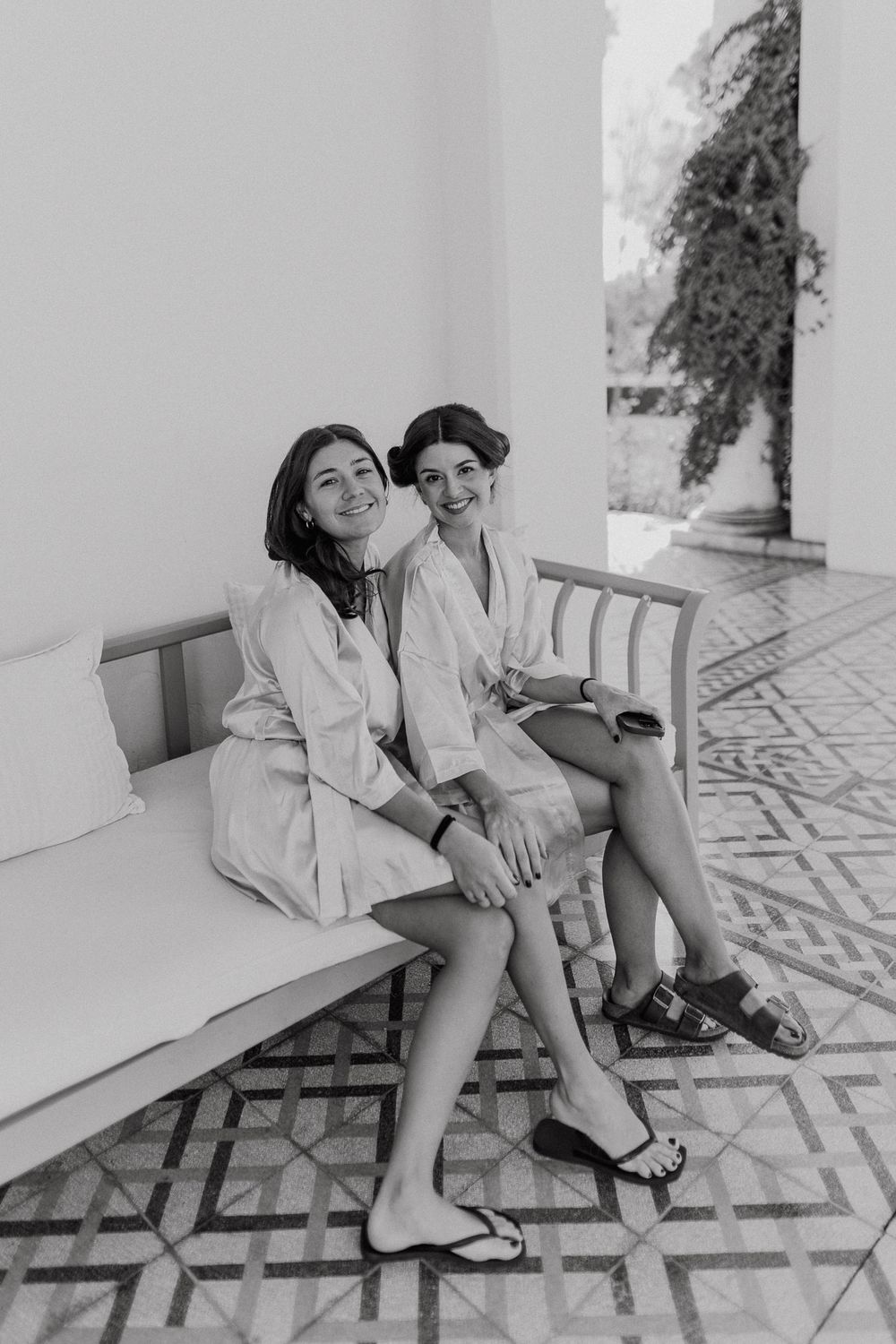 Two women in white robes sitting together on a bench, smiling at camera in black and white photo.