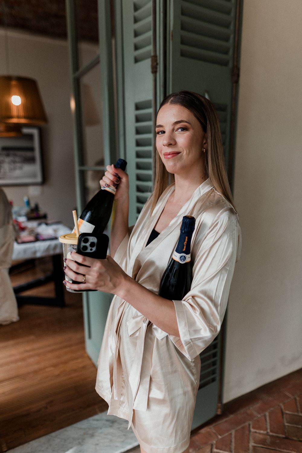 Young woman in silk robe holding champagne bottle and cocktail glass, smiling indoors near green shuttered doors.