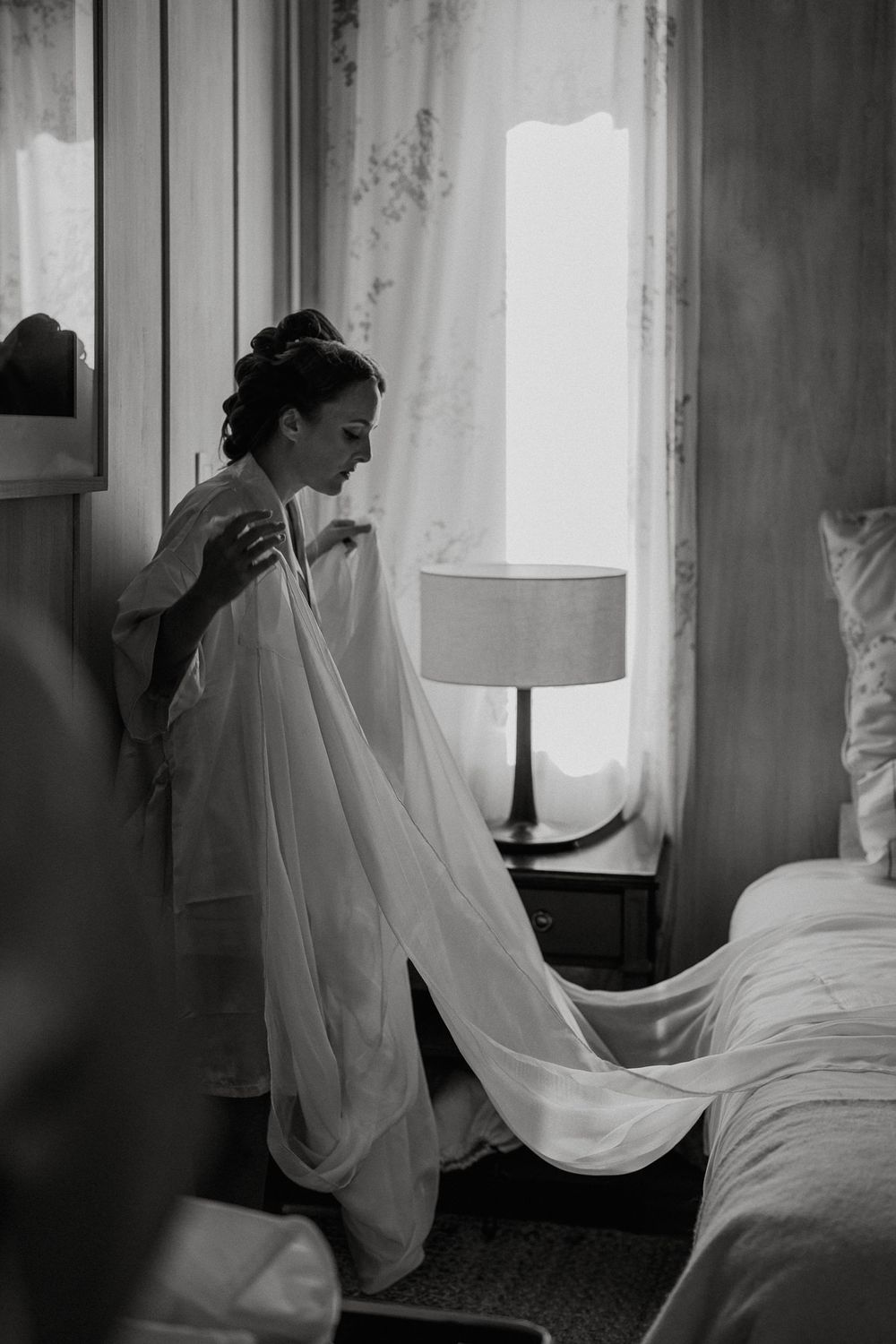 Black and white photo of a bride in a flowing white robe preparing by a window with soft natural light.