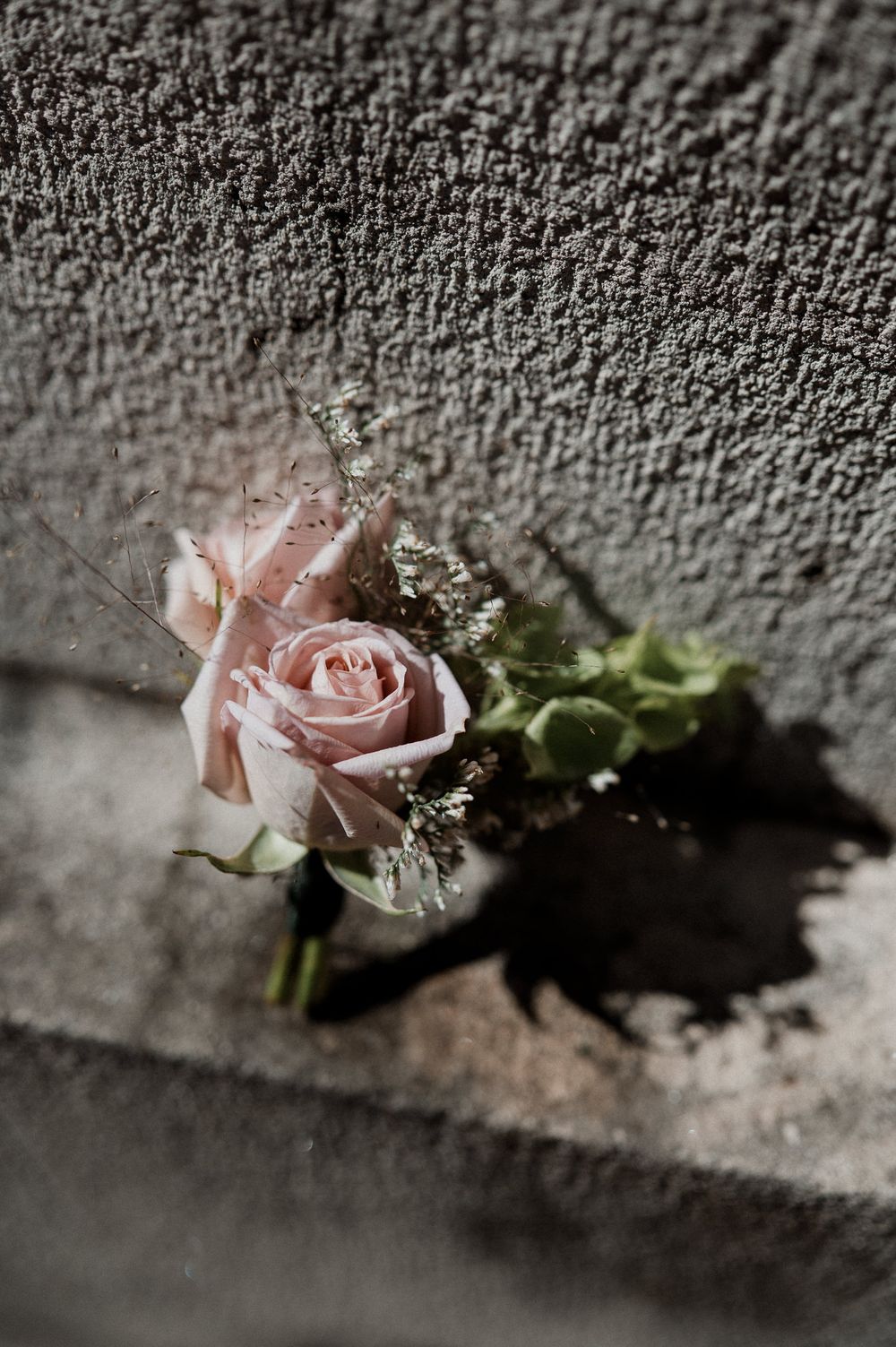 Small pink rose boutonniere with baby's breath resting against a textured gray concrete wall in soft natural light.
