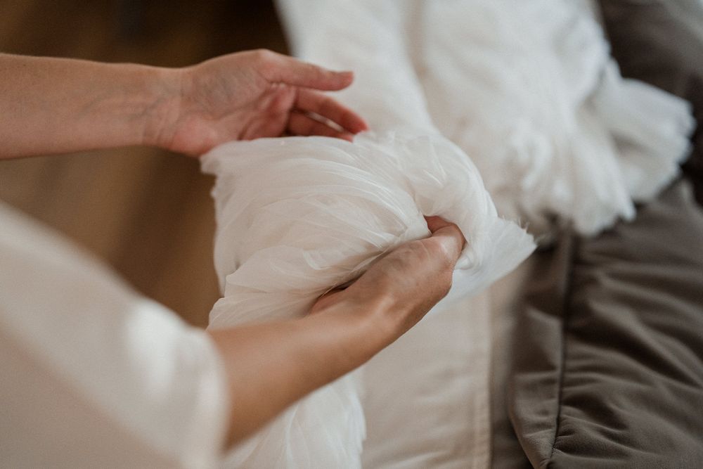 Hands carefully arranging white fabric pillow stuffing on a gray sofa during home bedding preparation.