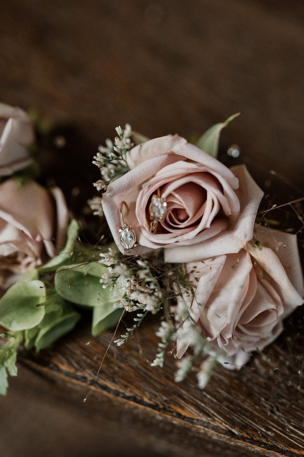 Dusty rose boutonniere with greenery and diamond wedding rings on rustic wood surface.