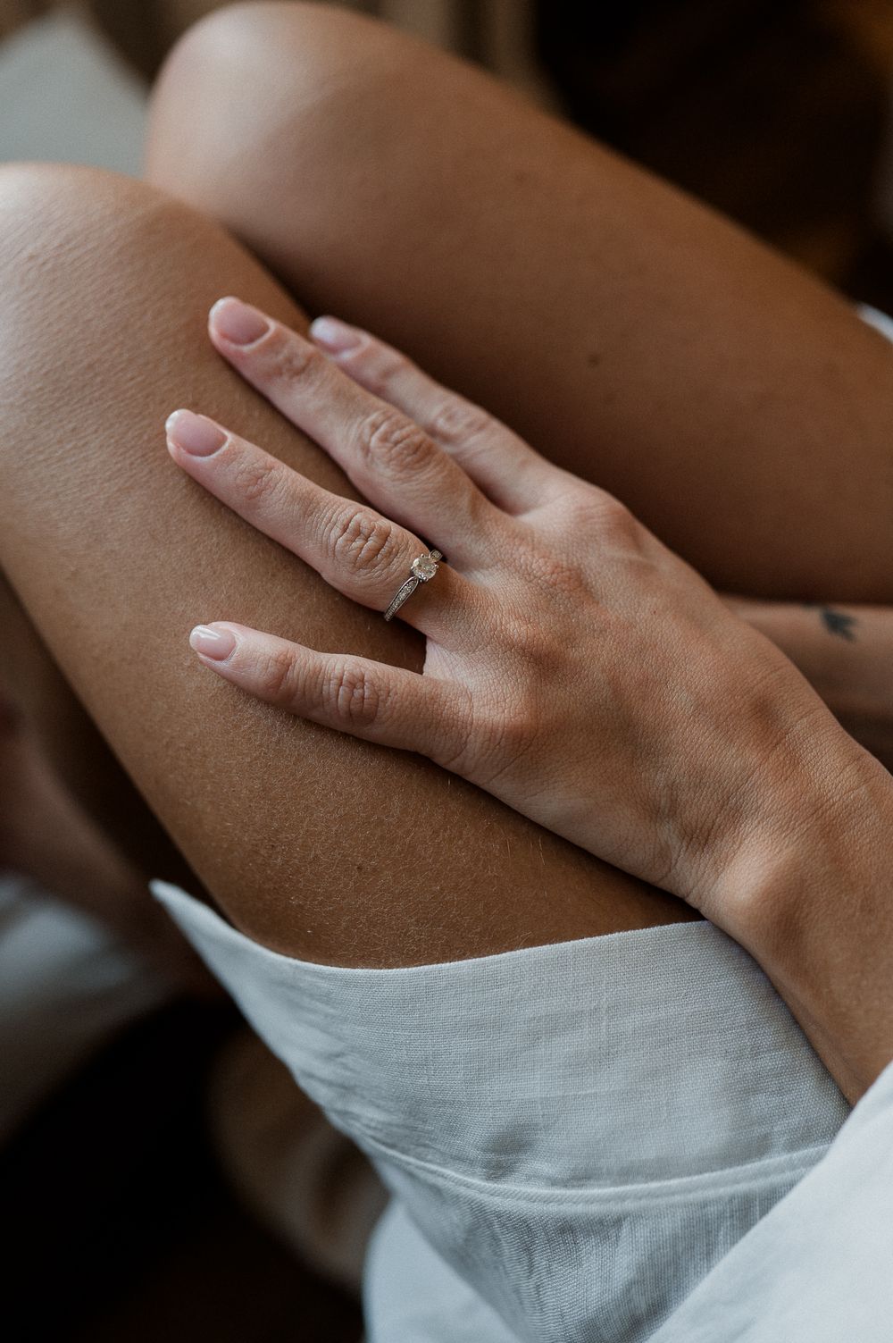 Close-up of a woman's hand with a diamond engagement ring resting on her knee, wearing a white dress.