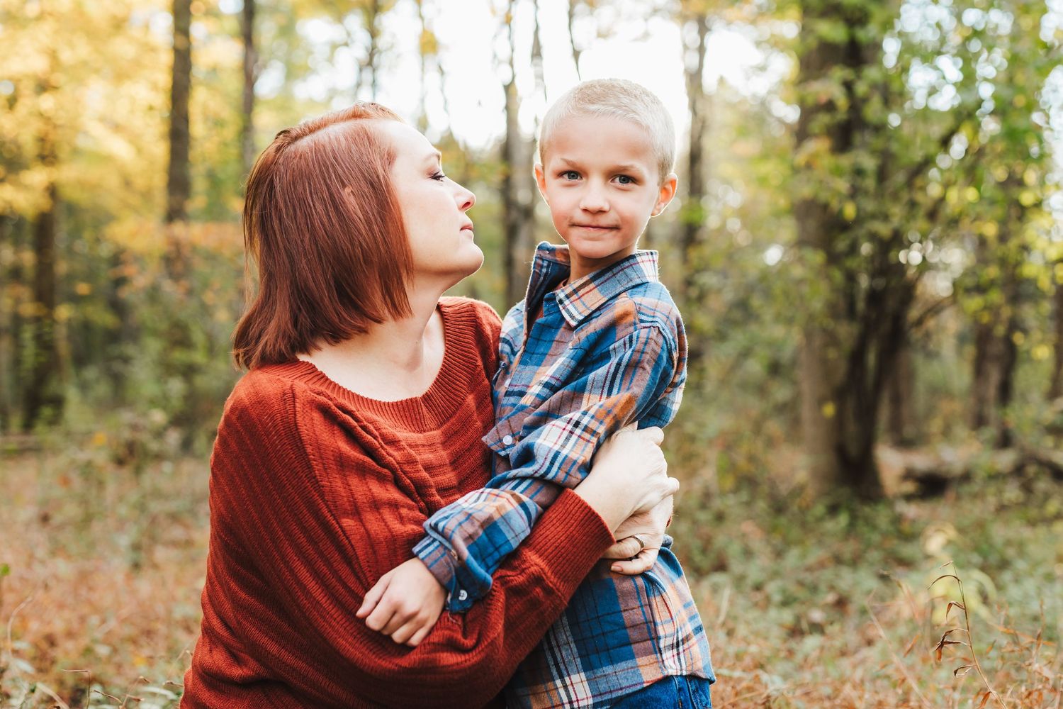 Family of three posing outdoors in autumn forest, parents and young son smiling among fall foliage in warm tones.