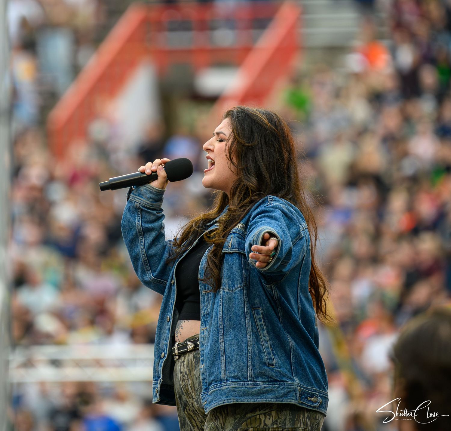 Young woman in denim jacket singing passionately into microphone on stage before large outdoor crowd.