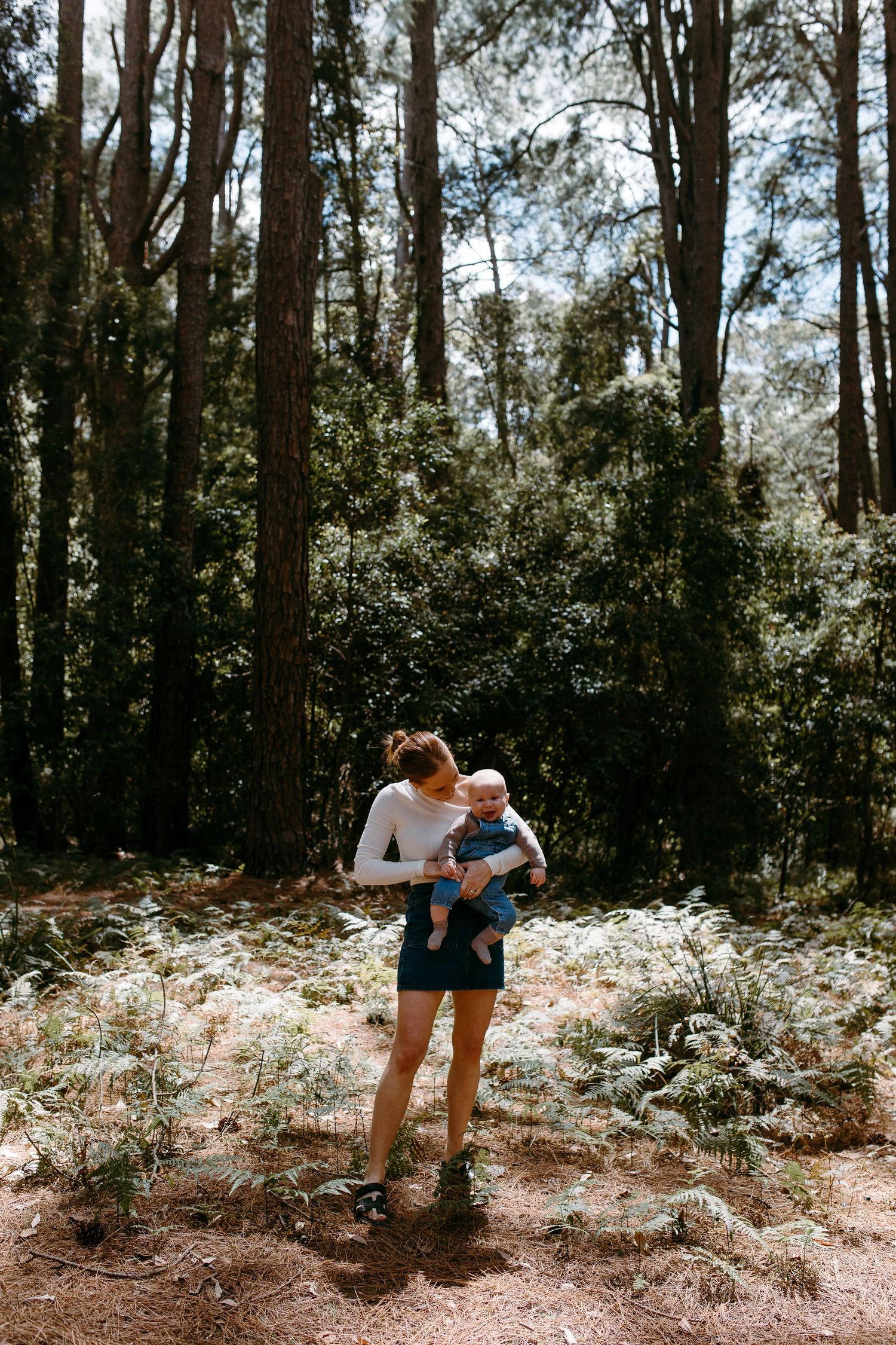 A young woman holds a baby in a sunlit forest clearing surrounded by ferns and tall pine trees.