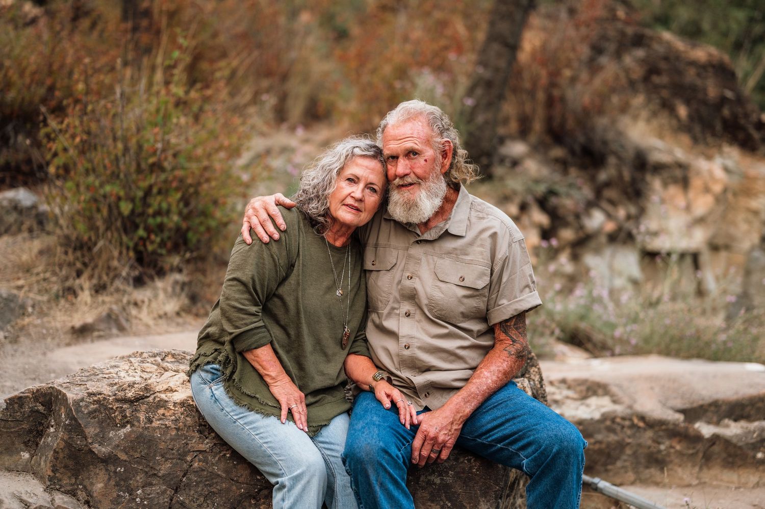 An elderly couple sits on a rock outdoors, embracing warmly with a natural hillside background.
