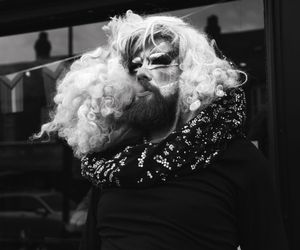 Black and white portrait of bearded person wearing white curly wig and sequined jacket.
