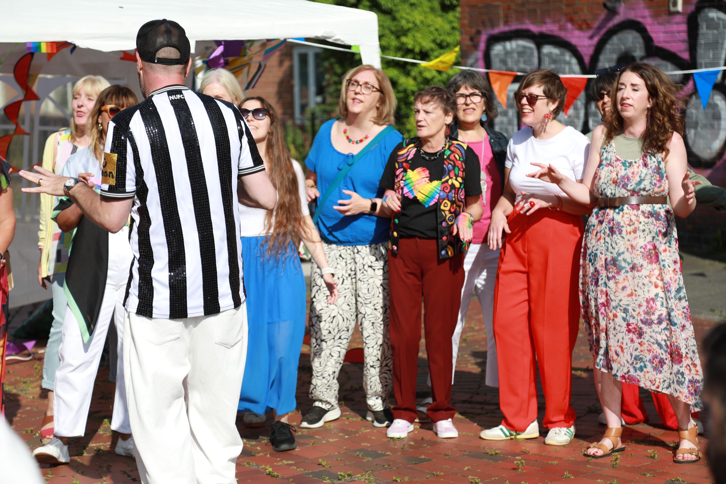 Group of people gathered outdoors at a community event near graffiti wall on sunny day.
