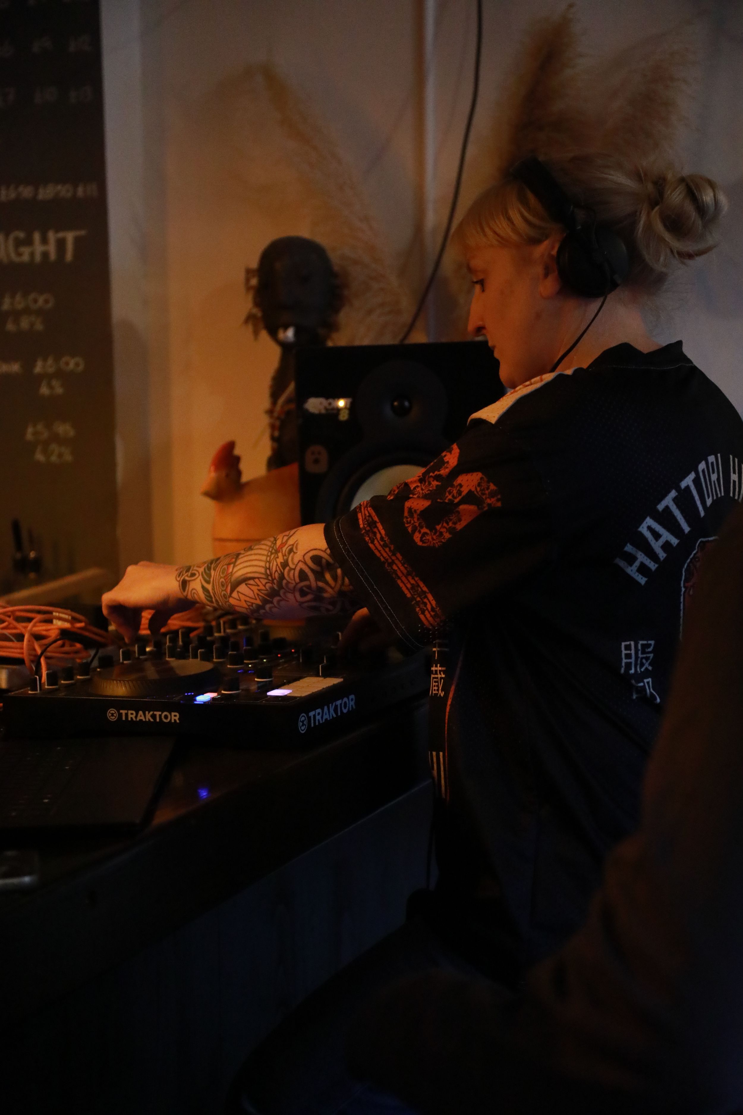 Female DJ with tattoos and headphones mixing on Traktor decks in a dimly lit venue.