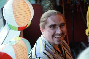 Smiling young person with blonde hair surrounded by colorful paper lanterns at an outdoor event.