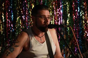 Tattooed musician playing guitar and singing into microphone against colorful sequin backdrop.