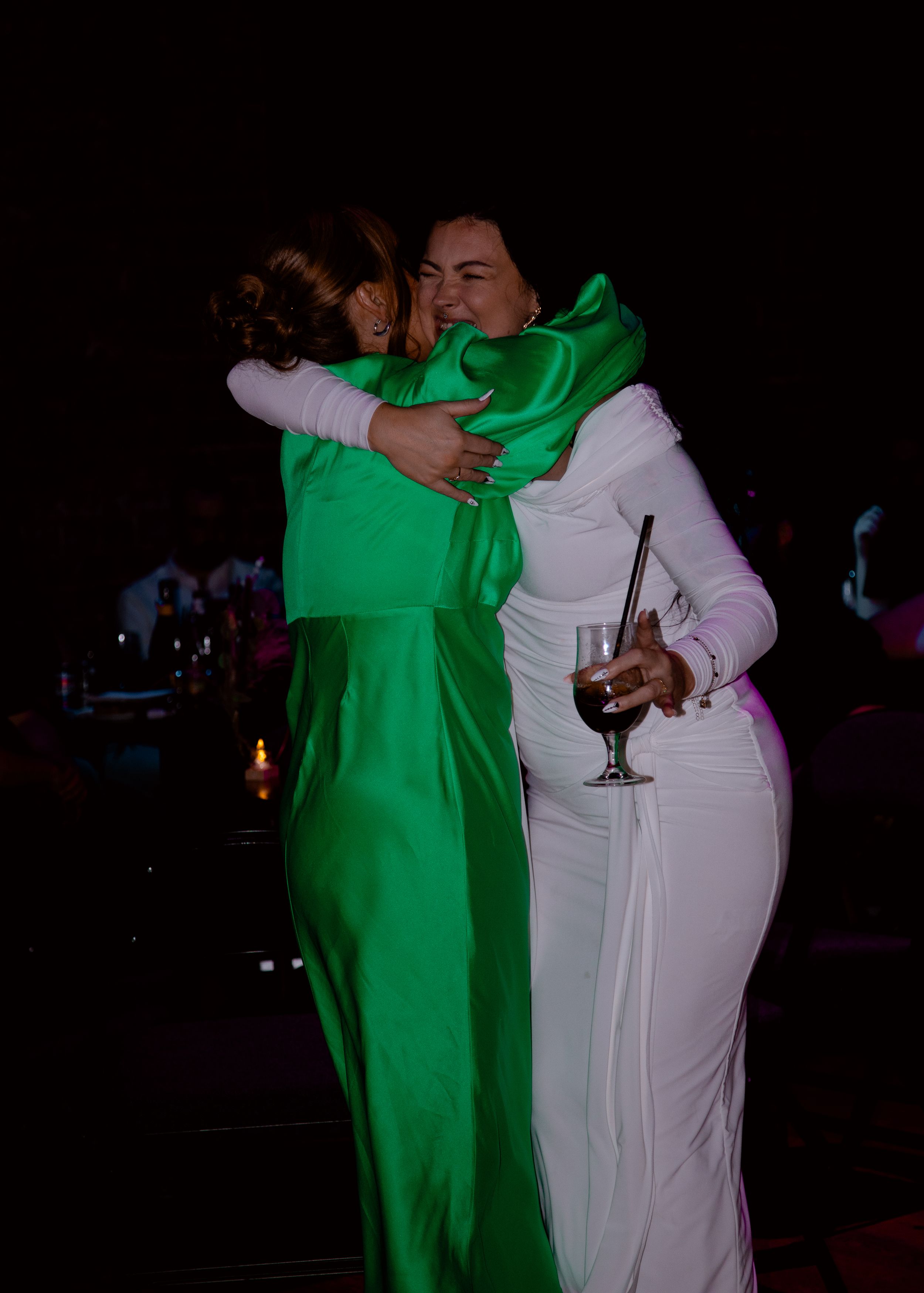 Two women hugging at a party, one in a green dress and one in a white dress.