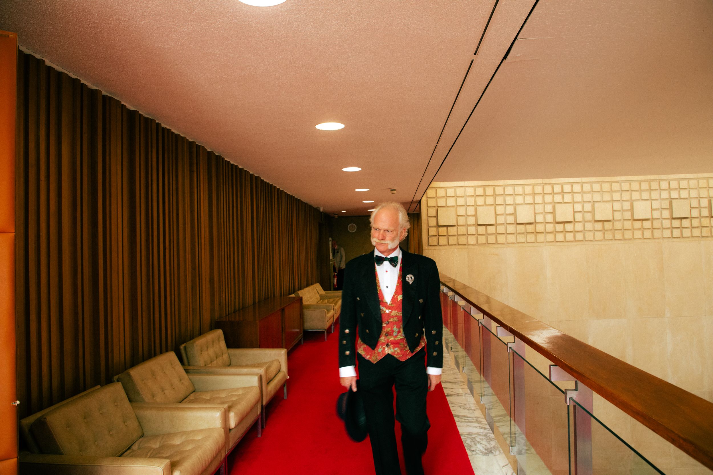 Elegant man in formal black tailcoat with red vest walking along a red carpet in a grand interior hallway.