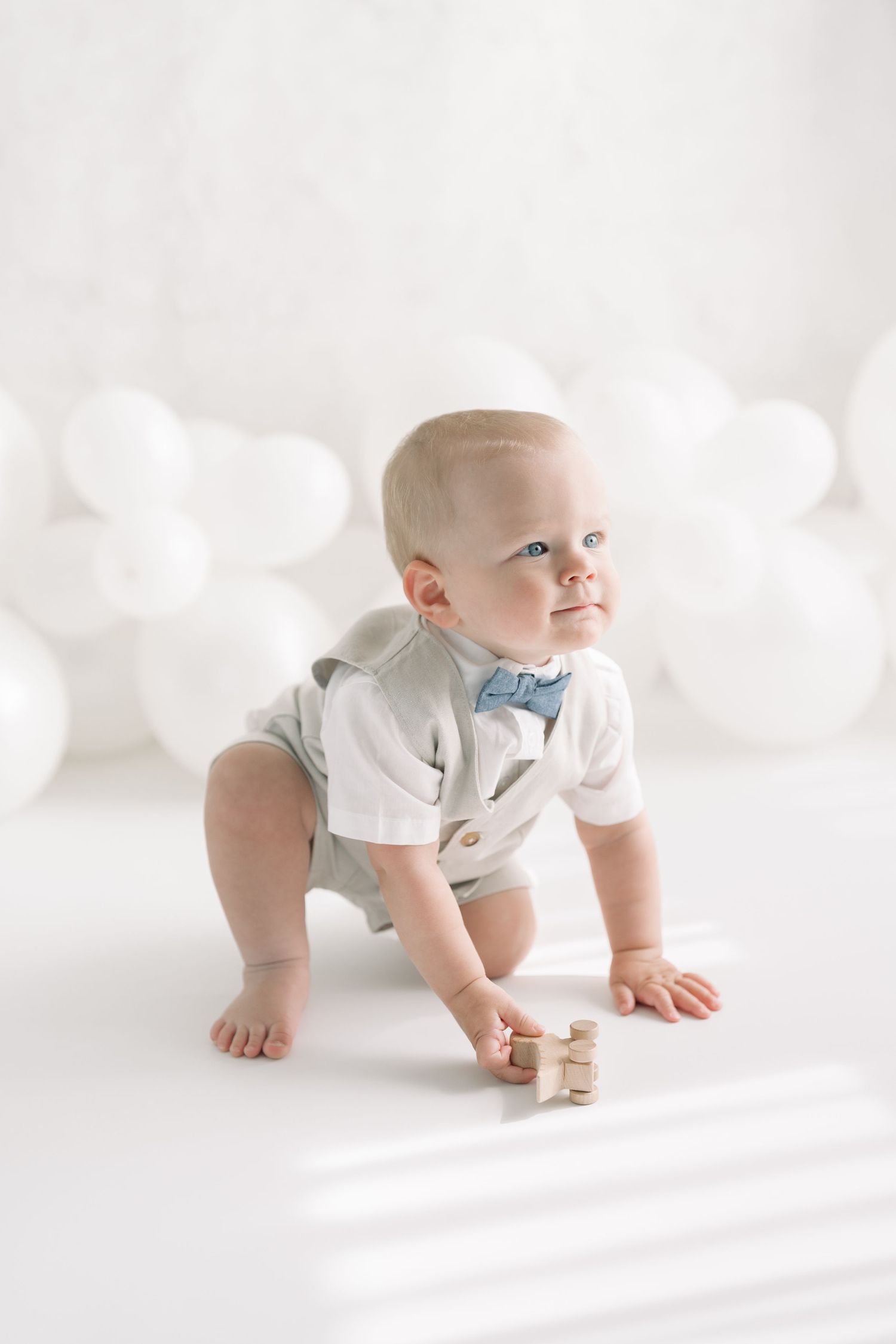 Bébé accroupi sur le sol blanc du studio dans le décor de ballons, un petit dinosaure bois naturel a la main, regardant vers ses parents