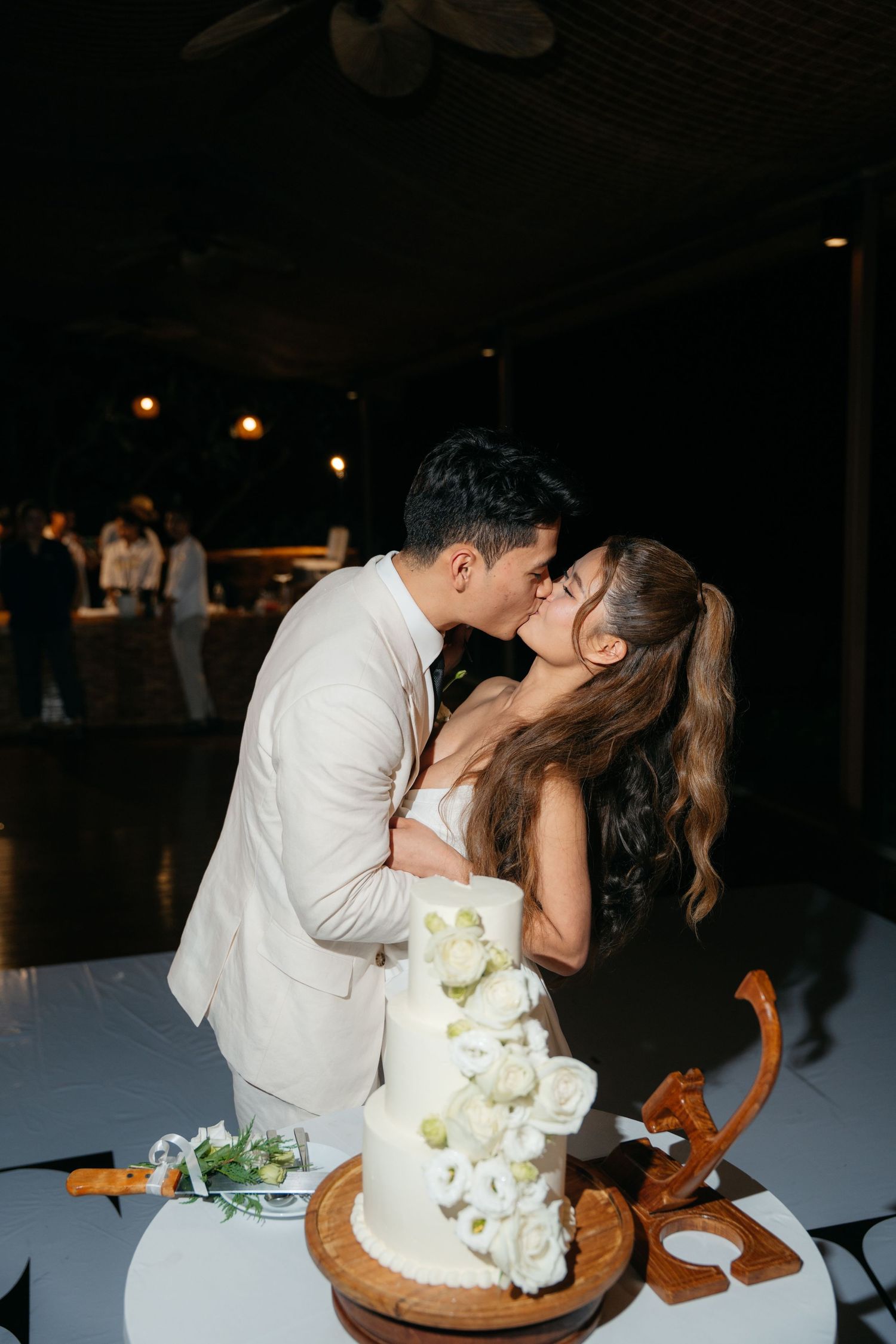 Newlyweds share a romantic kiss over a white floral wedding cake at their elegant indoor reception.