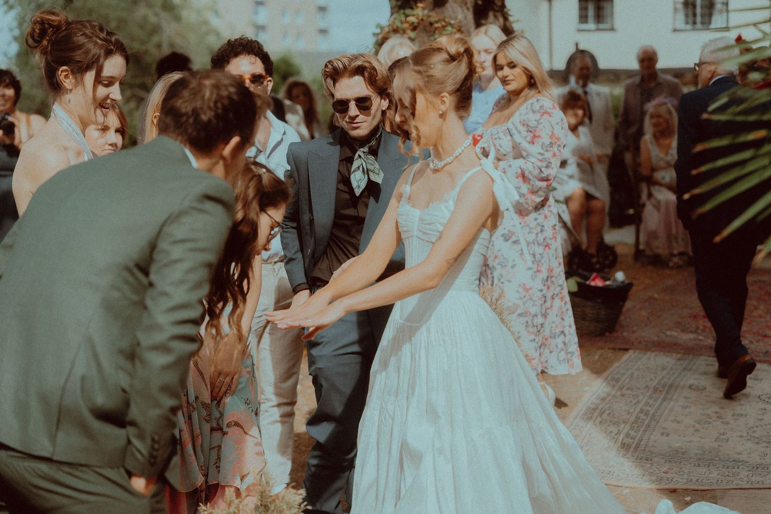 Bride in blue wedding gown and groom in green suit greet guests outdoors at a bohemian wedding ceremony.