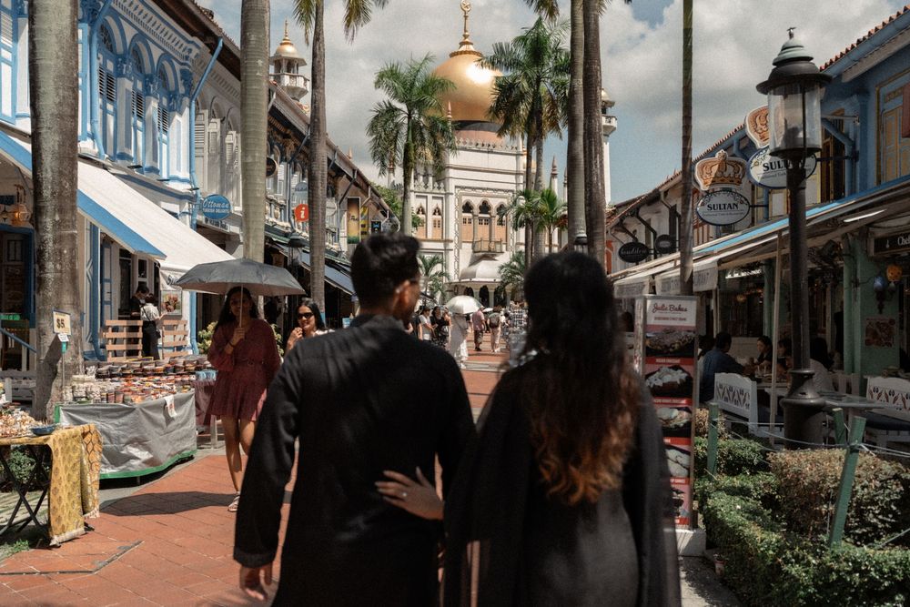 Couple walking hand in hand through a vibrant street market with palm trees and a golden domed mosque in Singapore.