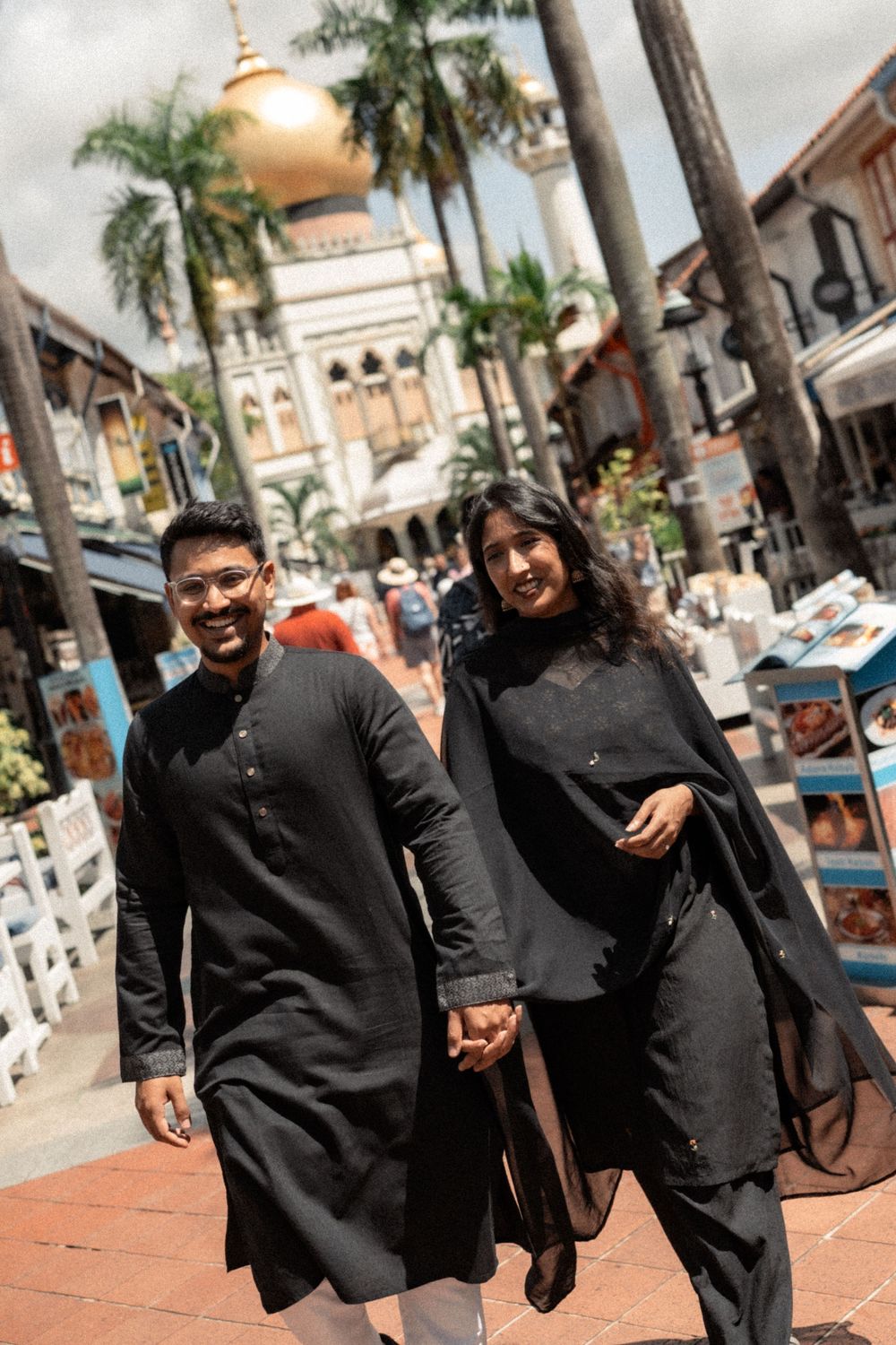 Couple in matching black traditional outfits holding hands near Sultan Mosque in Singapore's Arab Street.
