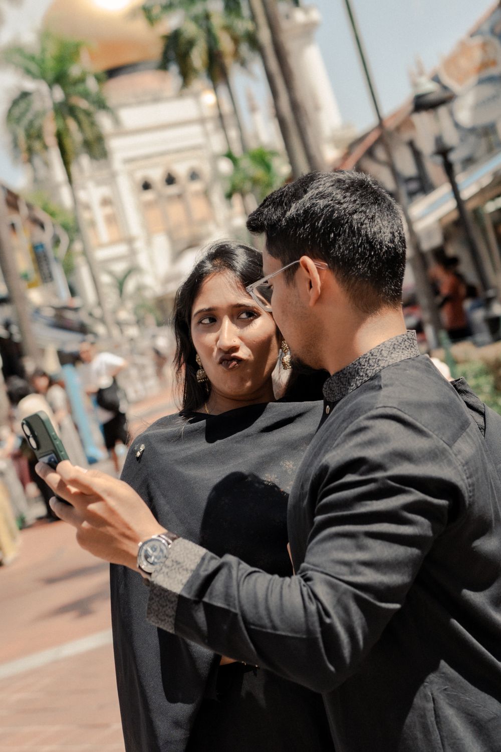 A couple in black outfits looking at a phone on a busy street with palm trees and a golden dome building.