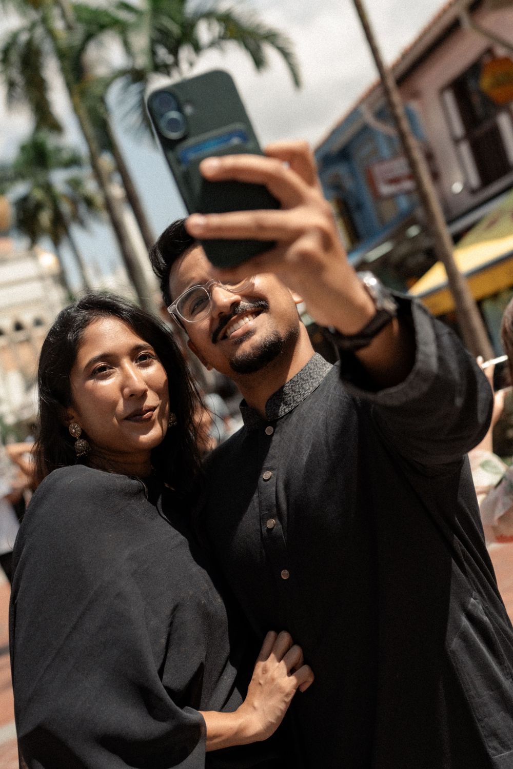 A couple dressed in black takes a selfie outdoors near palm trees and colorful buildings on a sunny day.