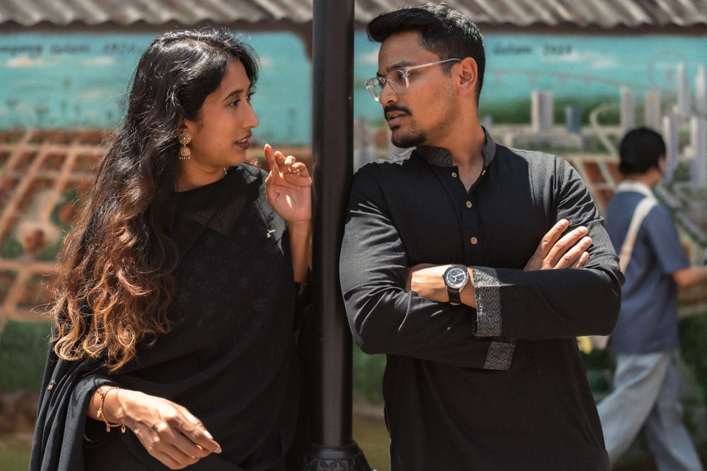 A man and woman dressed in black traditional attire engage in conversation outdoors near a decorative pole.