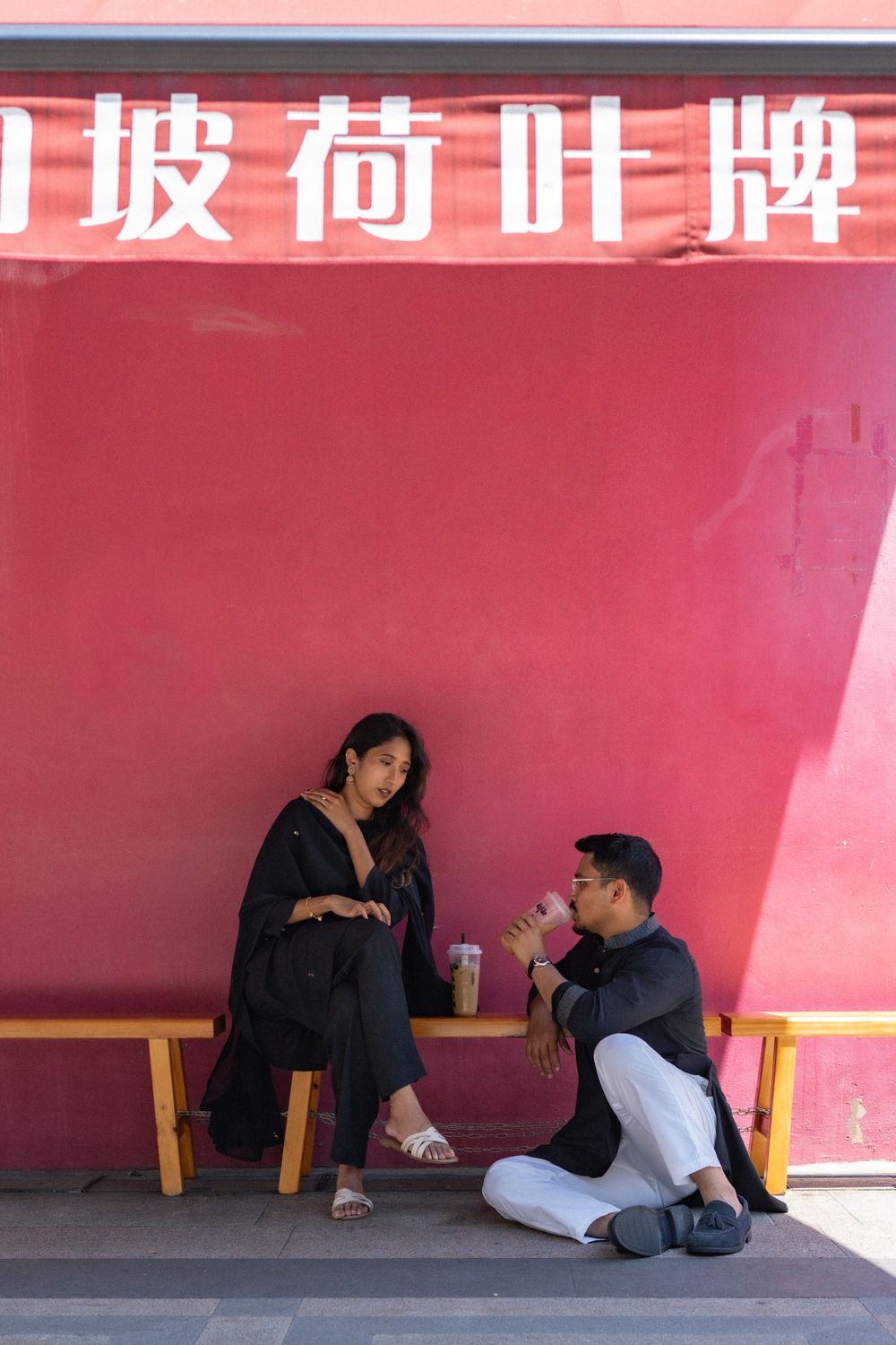 Couple relaxing at a red Chinese restaurant wall bench, woman in black outfit seated while man drinks bubble tea.