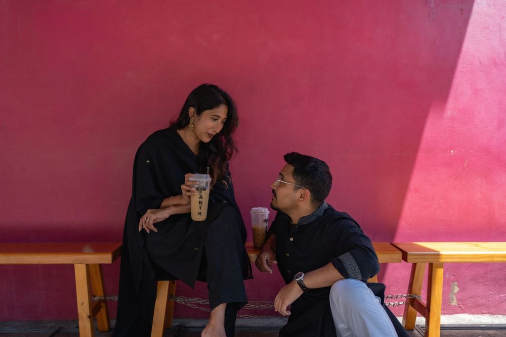 Young couple sharing drinks at an outdoor cafe against a bold pink wall, dressed in matching black outfits.