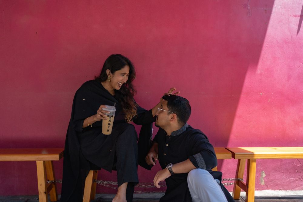 A couple in black outfits sharing a moment at a cafe, woman holding bubble tea against a pink wall.