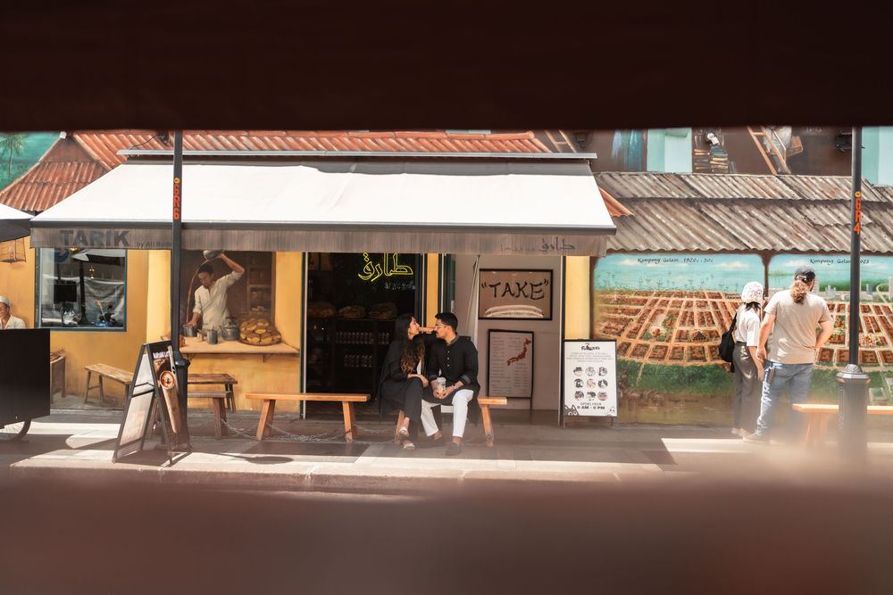 People sitting on a bench outside a cafe storefront with a mural on the wall in a sunny urban street.