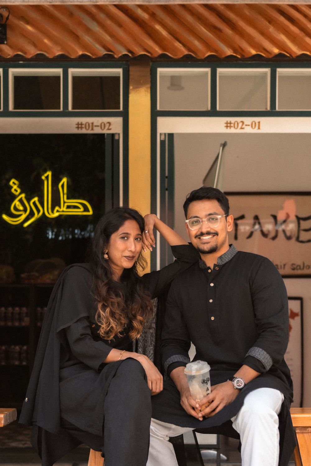 A young couple in black outfits sitting together outside a cafe with Arabic signage, smiling warmly.