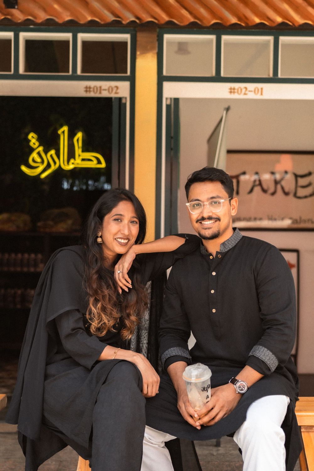 A smiling couple in matching black outfits sitting together outside a cafe with Arabic signage.