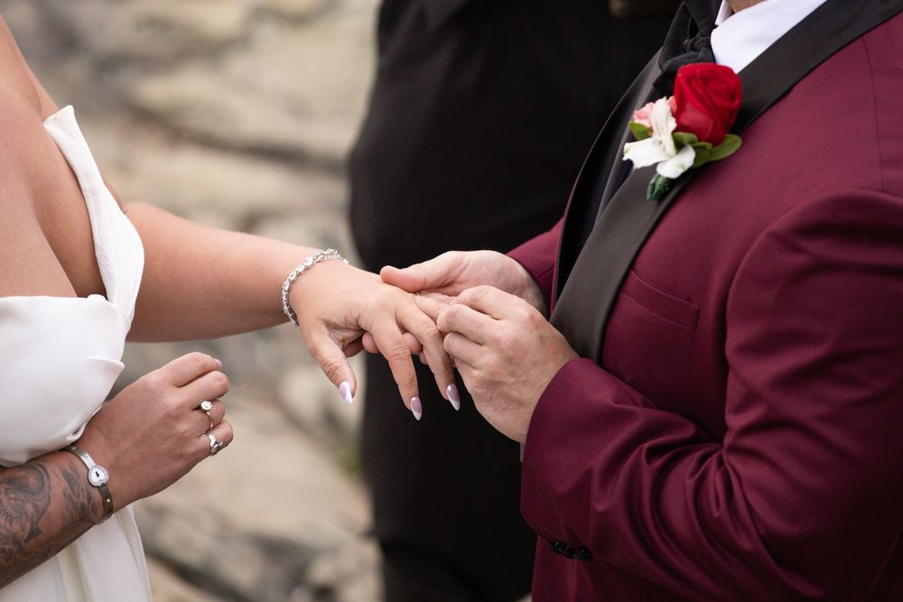 Groom in burgundy suit placing wedding ring on bride's finger during ceremony.