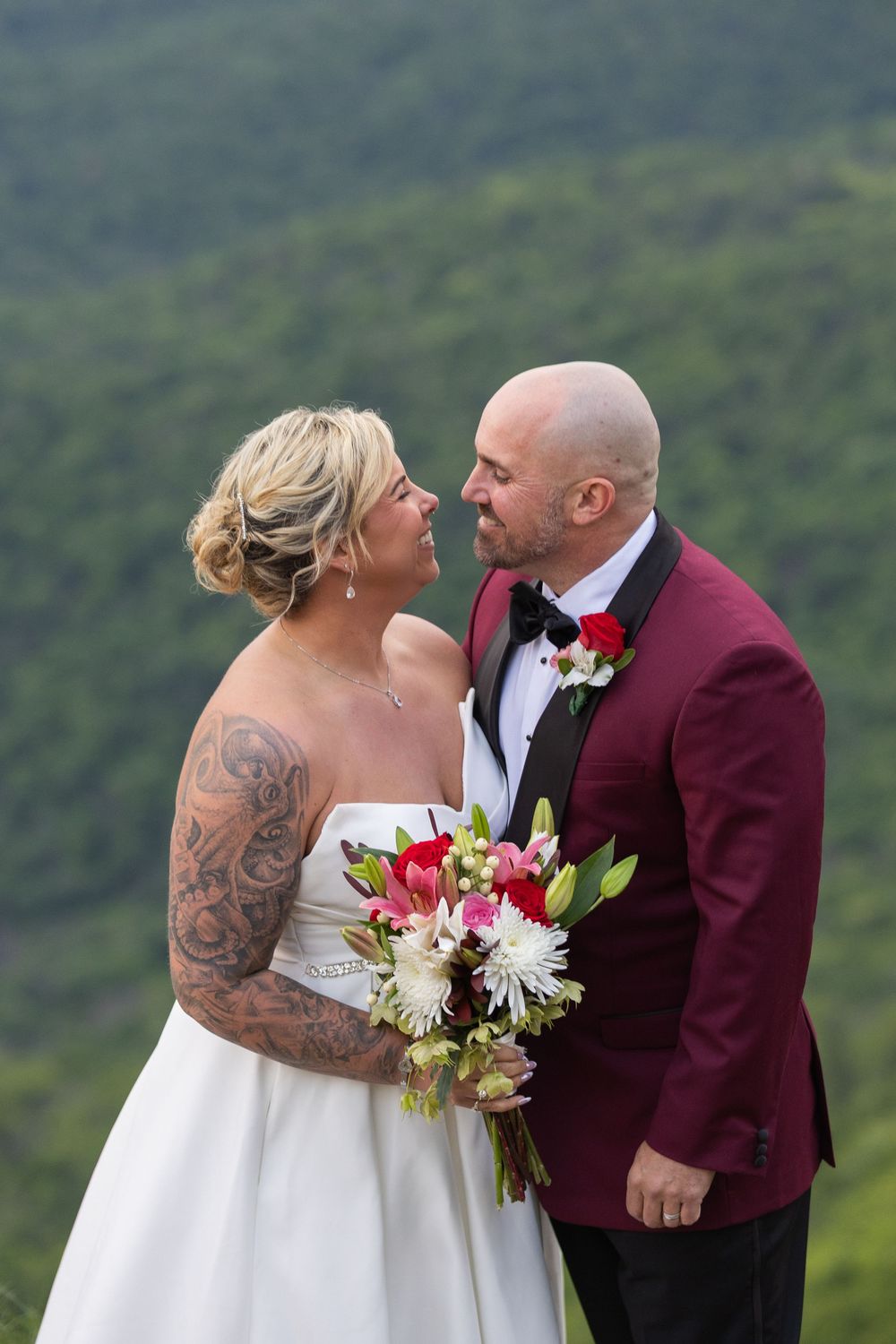 Bride with tattoos in white gown holds floral bouquet beside groom in burgundy tuxedo, sharing a romantic moment outdoors.