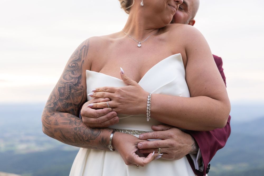 Bride in white strapless gown embraced by tattooed partner on mountain overlook at outdoor wedding ceremony.