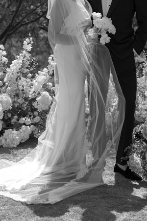 Wedding couple's lower bodies shown with flowing white dress and black tuxedo against garden backdrop with white flowers.